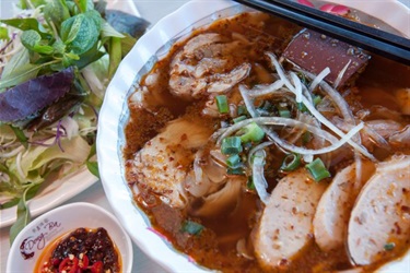 A large bowl of Bun Bo Hue spicy Noodle soup, plate of purple & white cabbage herbs and sate chili dipping sauce.