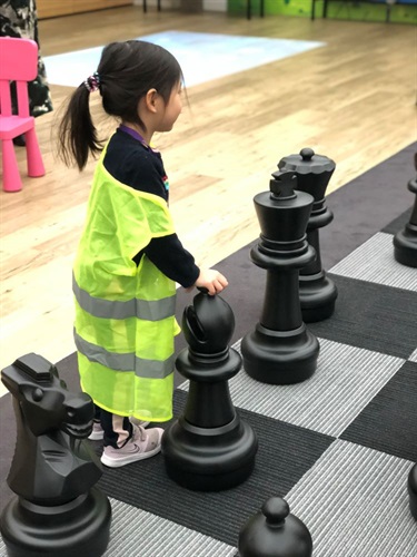 Young girl wearing high visibility vest standing on giant chess board while holding a black bishop piece