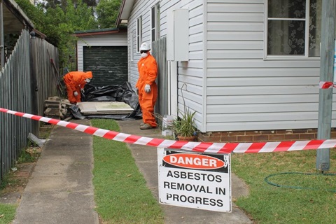 People in hazard suits behind a line of safety tape with sign saying Danger Asbestos removal in progress