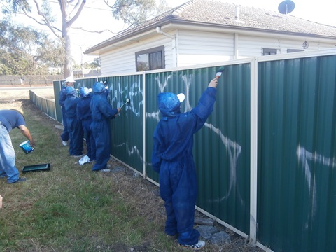 Group of people wearing blue protective suits while painting over graffiti on fence 