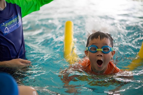 Young boy wearing swimming goggles using a pool noodle to swim