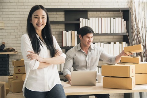 A young woman smiling and posing with her arms crossed while sitting on a desk. Behind the desk, a young man is looking at a laptop while arranging cardboard boxes. 